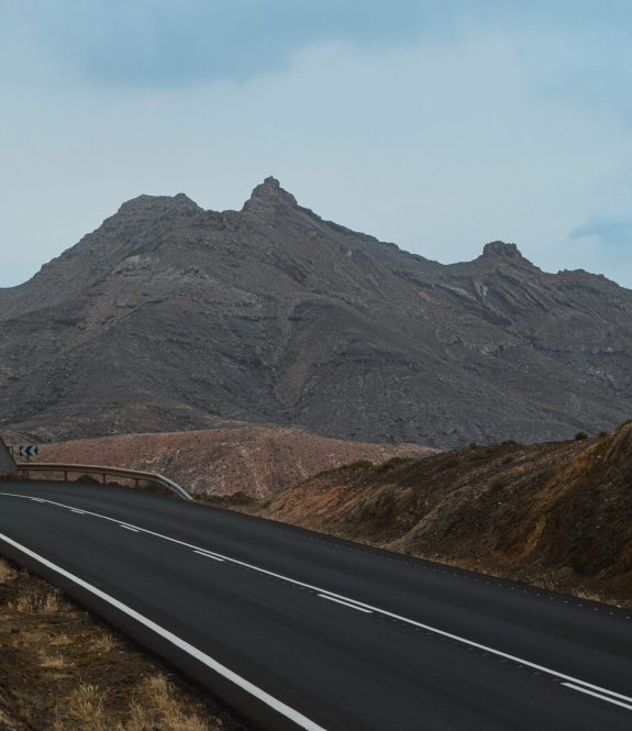 This image depicts a desolate, winding road leading towards a rugged mountain range. The road, with its smooth, dark asphalt and white lane markings, cuts through a barren, rocky landscape with minimal vegetation. The surrounding hills are composed of dry, brown earth, creating a stark, almost otherworldly scene. In the background, the towering mountain has a jagged, angular peak that dominates the horizon under a cloudy, pale blue sky. The road curves sharply to the left, with a triangular road sign visible at the bend, emphasizing the remote and solitary nature of this location
