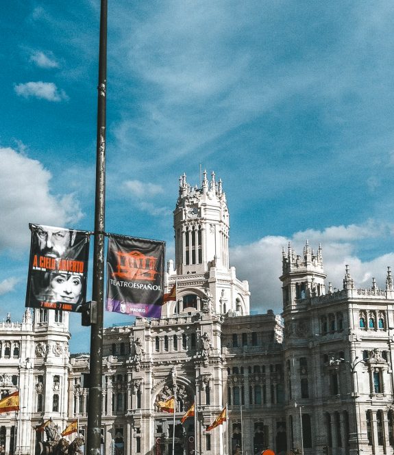 This image features a grand, ornate building with a stunningly detailed facade, topped by several towers and spires. The architecture is a striking example of neoclassical or Gothic Revival style, with intricate stonework and decorative elements. The building is likely a historic or government structure, standing tall against a backdrop of a bright blue sky with scattered clouds. Spanish flags are visible around the building, adding a sense of national pride and indicating the location is likely in Spain, possibly in a major city like Madrid. The overall scene is vibrant and bustling, capturing both the architectural beauty and cultural significance of the area.