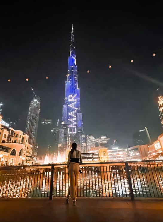 Nighttime view of the Burj Khalifa and Dubai Fountain in Downtown Dubai, with a woman admiring the illuminated skyline. The iconic Burj Khalifa, the tallest building in the world, lights up with vibrant displays, creating a mesmerizing backdrop for visitors. Surrounded by beautifully lit traditional Arabian architecture and bustling city lights, this location is a must-visit for Dubai travelers looking to experience the city's luxury and modernity. Perfect for nightlife, shopping, and sightseeing
